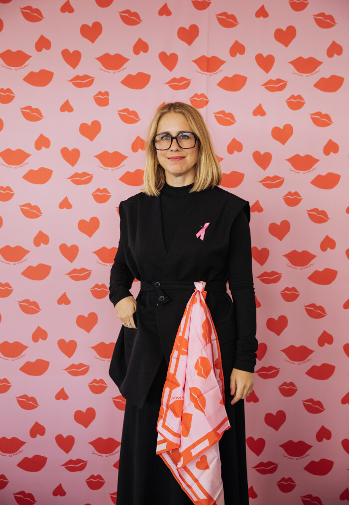 SCARF FOR SCARS X CARIN WESTER. Designern Carin Wester med rosa bandet på bröstet och en scarf som hon designat till förmån för bröstcancerforskningen. Scarfen är i 100% siden och 70 x 70 centimeter stor. Den är rosa med röda läppar och hjärtan på. Säljs exklusivt på Åhlens och ahlens.se och 100 kronor på varje såld scarf går till forskningen.