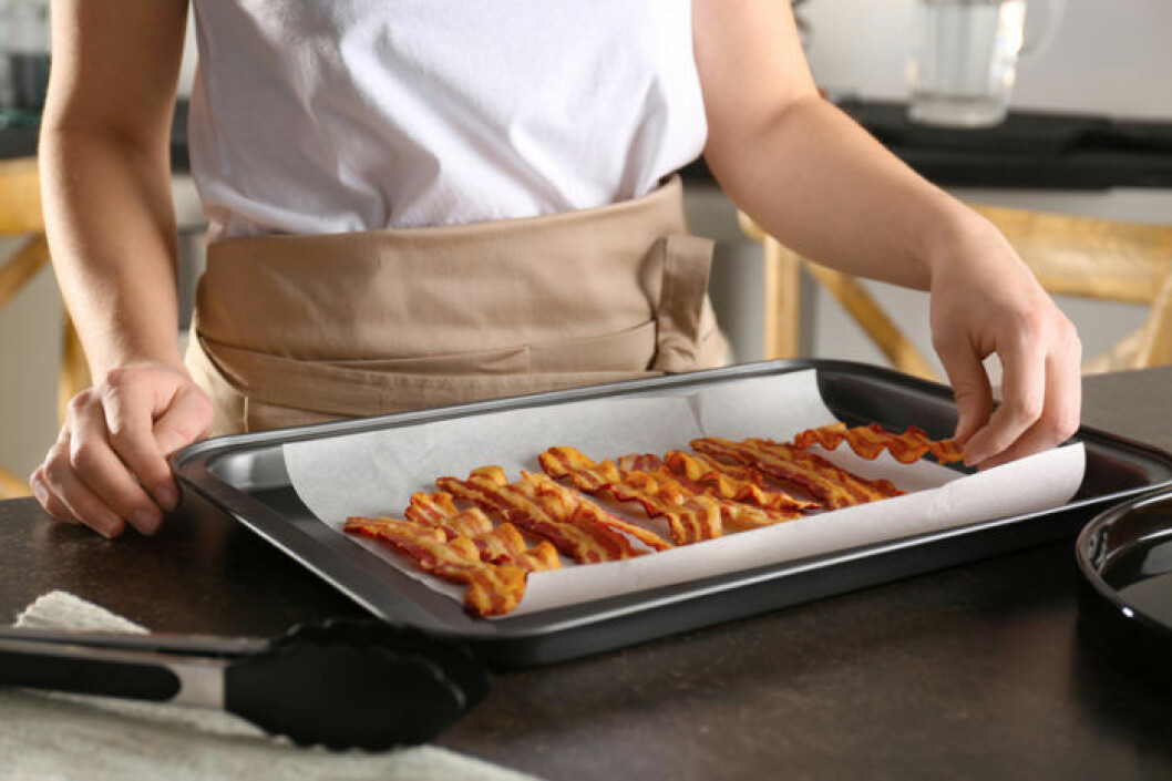 Tillaga bacon i ugnen för mindre kladd. Bild: Shutterstock/IBL Kvinna med plåt med bacon tillagad i ugn.