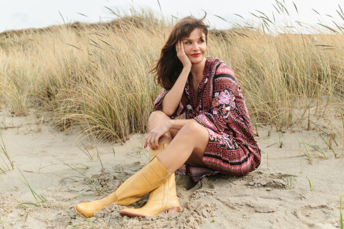 Helena Christensen sittandes i sanden på en strand med vass i bakgrunden. Hon har en kort tunika-klänning med uppvikta ärmar. Klänningen är mönstrad i rosa, rött, vitt och svart. Till det har hon brun-stövlar som slutar halvvägs upp på vaden. Klänning från Odd Molly.