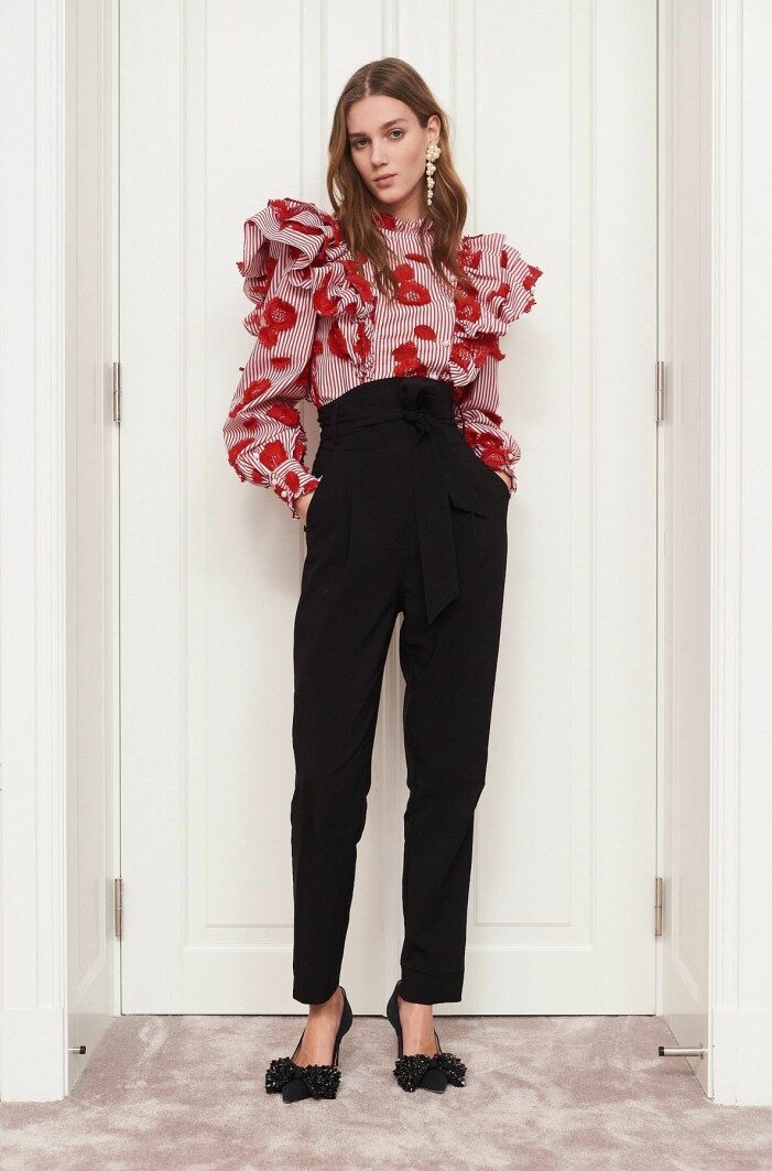 Blus, Custommade. Modell med svarta kostymbyxor med hög midja och svarta pumps med rosett detalj. På överkroppen har hon en röd och vitrandig blus med volang vid hals och stora volanger vid axlarna. I öronen har hon långa pärlörhängen. Blus från Custommade.