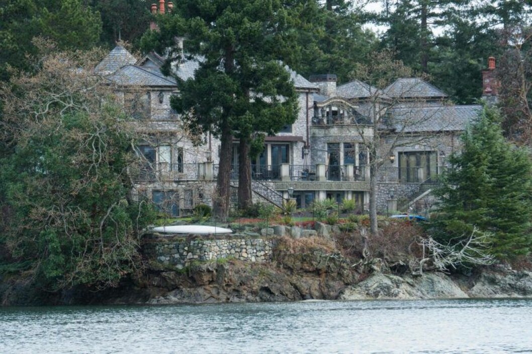 Harry och Meghans nya hus på Vancouver Island, Kanada. Ett stort grått hus i sten som ligger dolt bland träd och med vatten framför