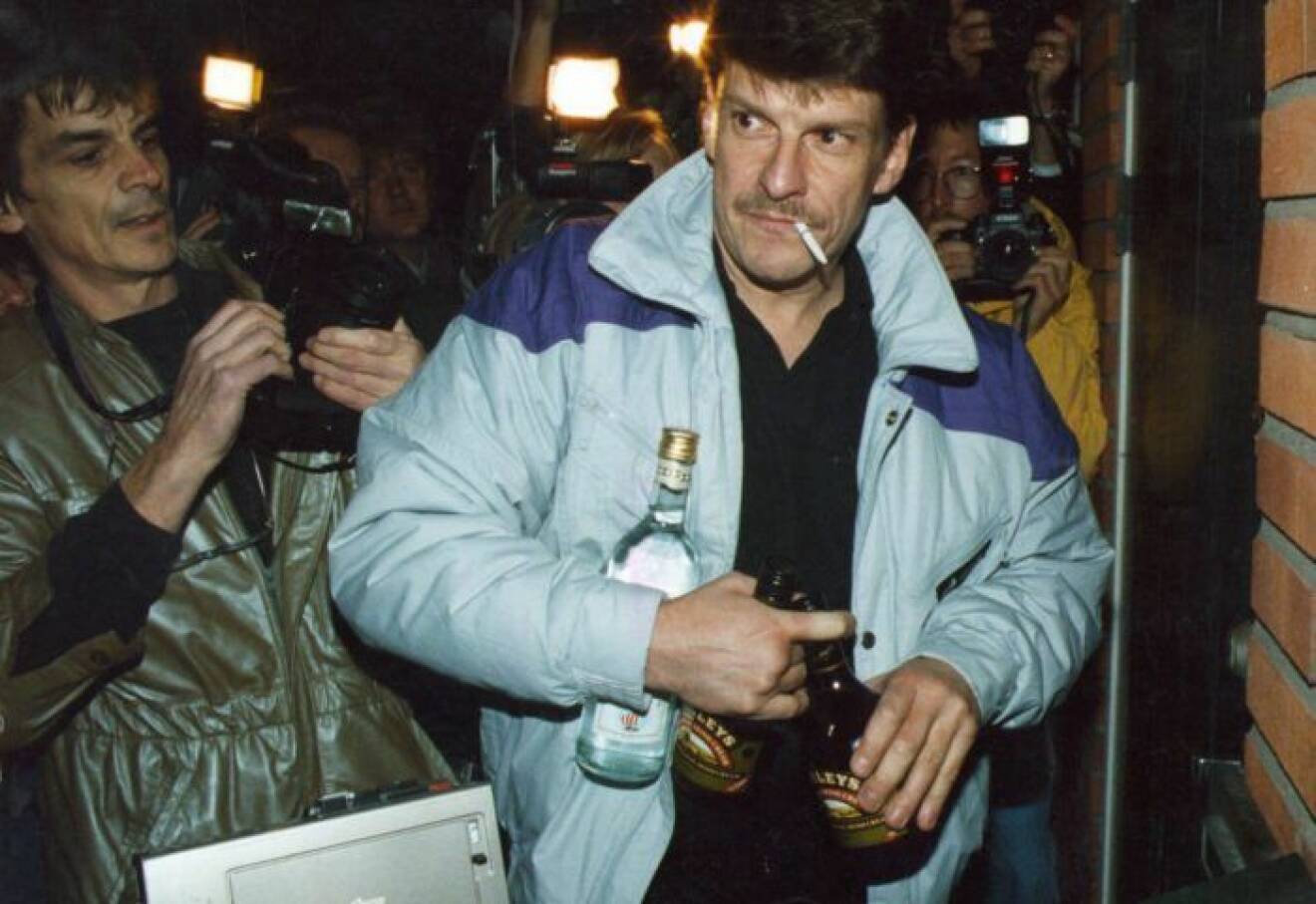 En man har ljusblå jacka på sig och en cigarett i mungipan. I handen har han flera flaskor med alkohol