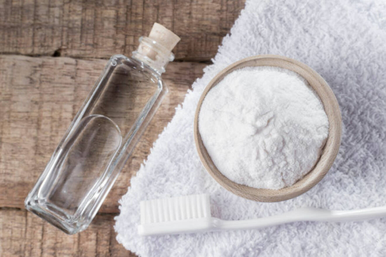 En flaska ättika bredvid en skål med bakpulver som står på en vit handduk. På Handduken ligger även en tandborste.