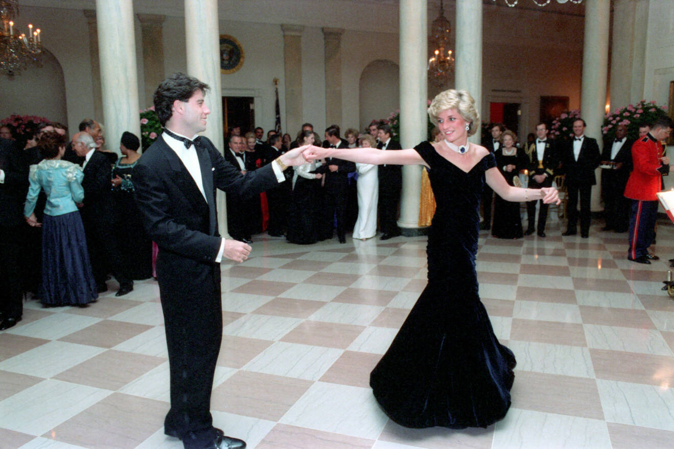 Prinsessan Diana dansar med skådespelaren John Travolta. Hon bär en svart långklänning med bara axlar och ett brett pärlhalsband. John har svart smoking.