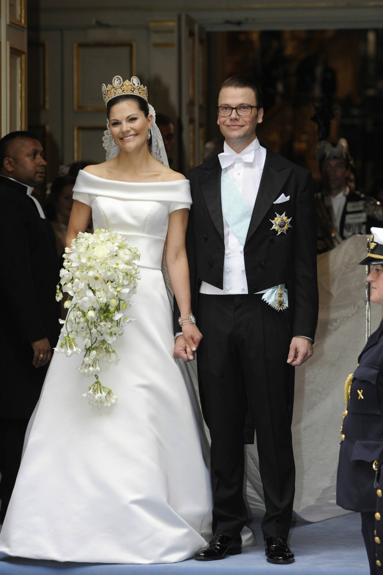 Victoria och Daniel precis efter vigselcermonin. Victoria har en krona, sin vita, minimalistiska bröllopsklänning med båtringning, en blombukett med vita blommor. Daniel har frack.