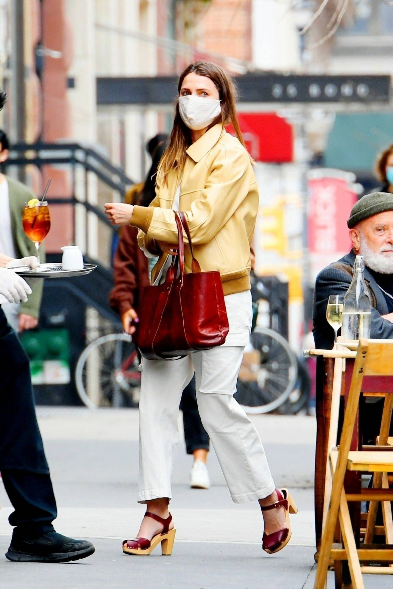 Skådespelerskan Keri Russel i burgundy-färgade träsandaler. Hon matchar med väska i samma nyans, vita, croppade byxor med ficka på låret, ljusgul skinnjacka och över ansiktet bär hon ett vitt munskydd.