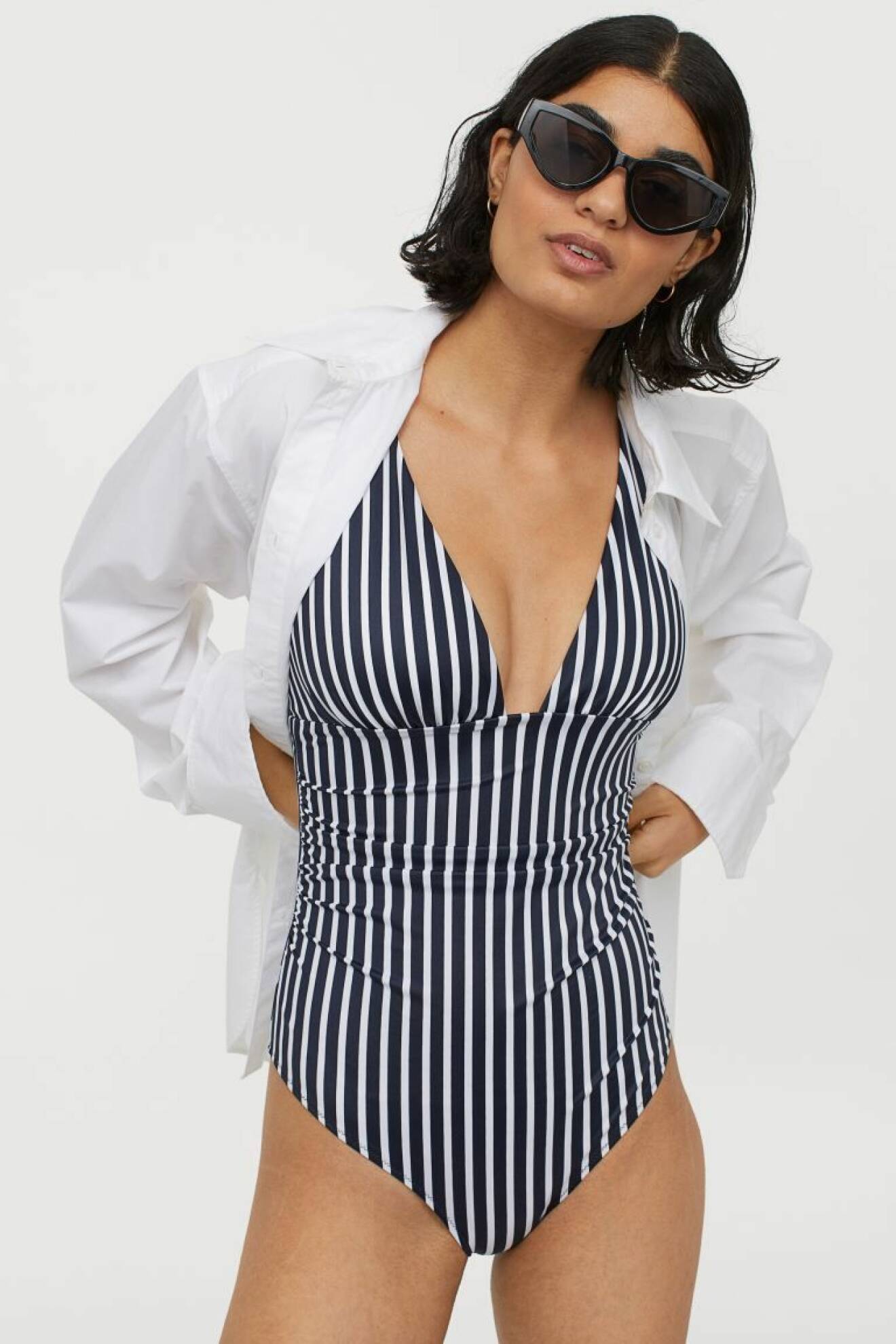 Modell med randig baddräkt i blått och vitt. Över har hon en vit skjorta och mörka solglasögon. Baddräkt från H&amp;M.