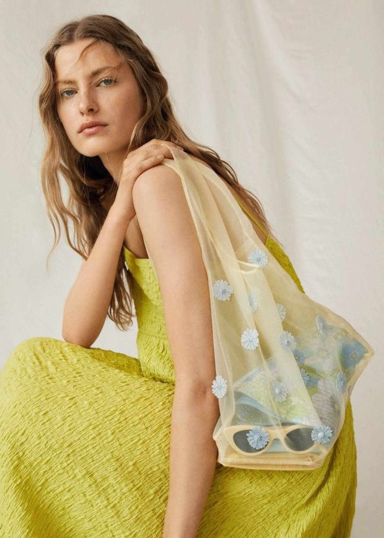 Modell med knallgul klänning och en ljusgul väska i påsmodell över axeln. Transparent väska i organza med ljusblå blommor i 3D på. Väska från Mango.
