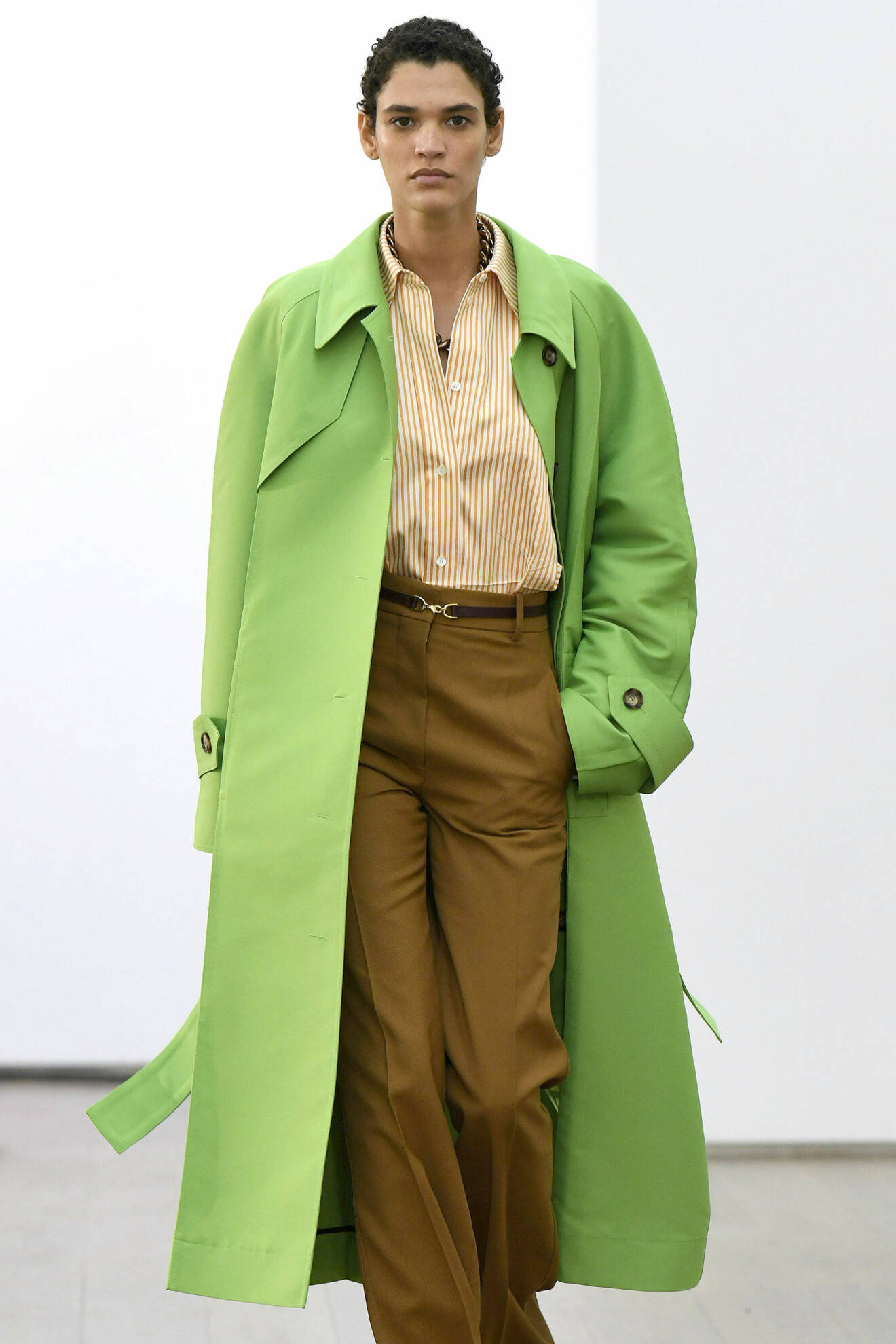 Modell i gul och vit randig skjorta, bruna byxor i dressad, oversizad modell och en grön trenchcoat över. Look från Victoria Beckham ss21.