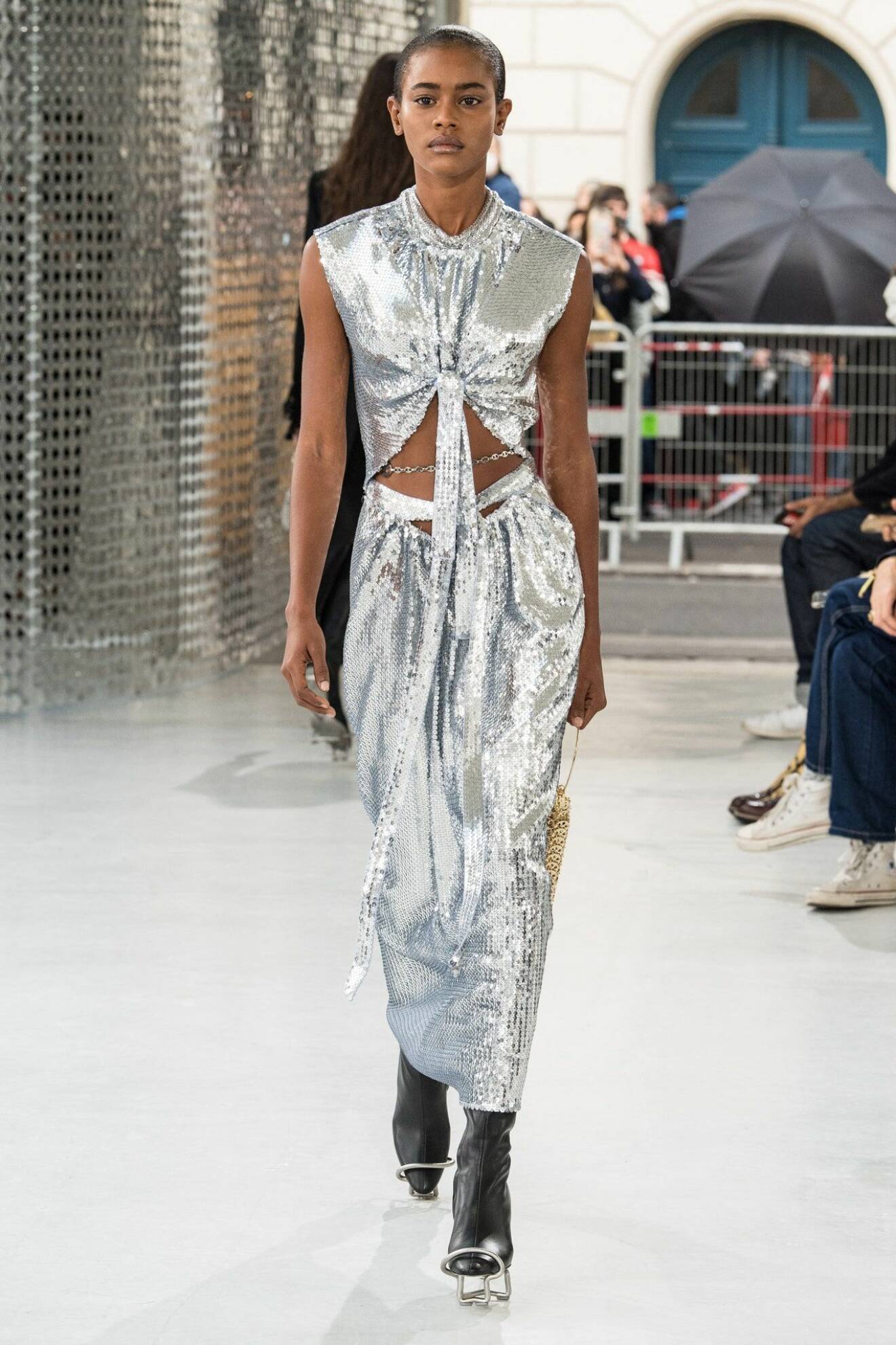 Modell med midilång klänning i silverfärgade paljetter. Stor cutout vid magen och knytdetalj som hänger ned från bystet. Look från Paco Rabanne ss21.