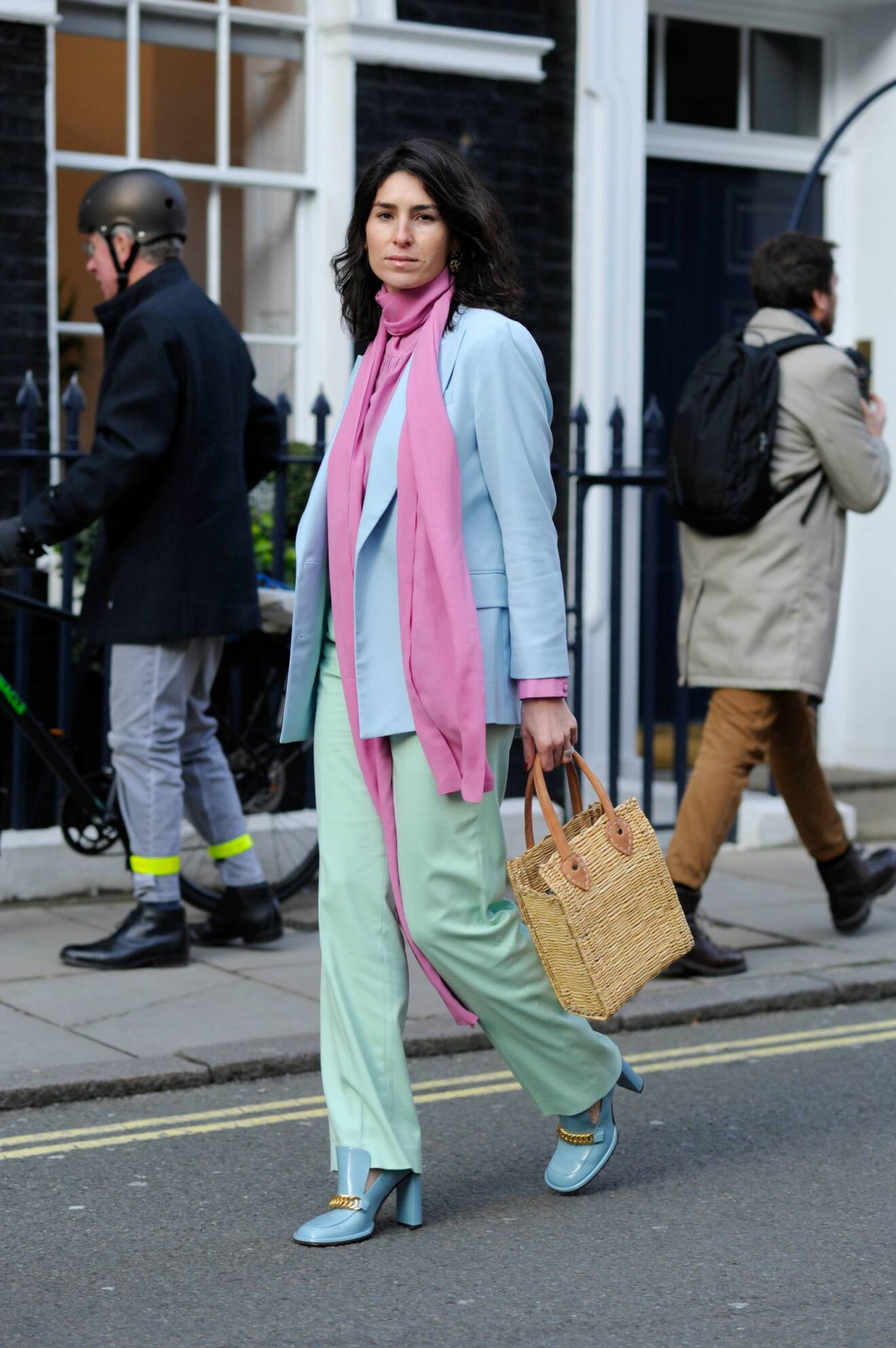 Kvinna i pastellfärgad outfit. Rosa, lång scarf, ljusblå blazer, gröna byxor och blå loafers med klack och guldfärgad detalj vid tån.