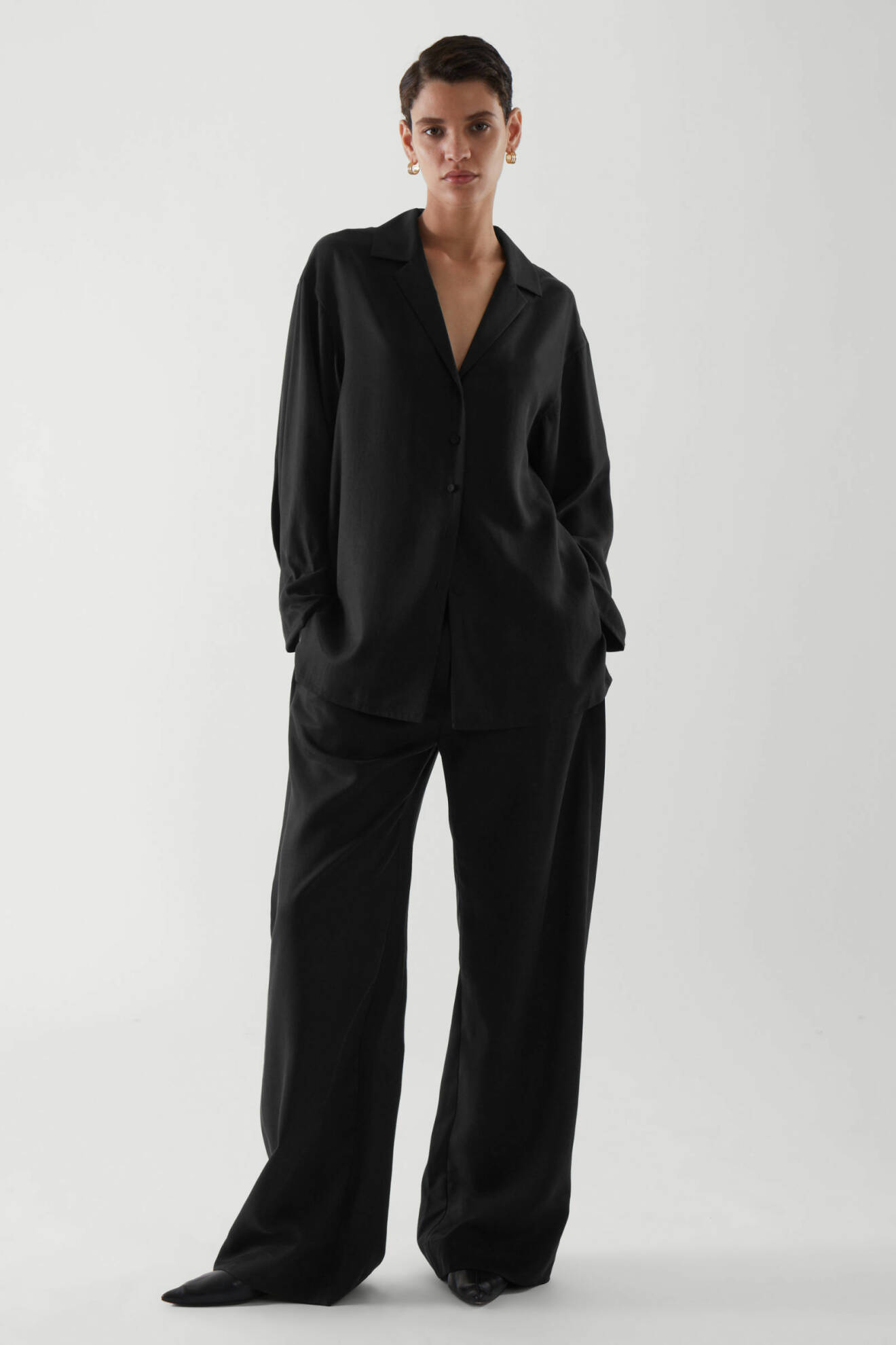 Modell med svart, minimalistisk skjorta och svarta, långa byxor i rak modell. Outfit från Cos.