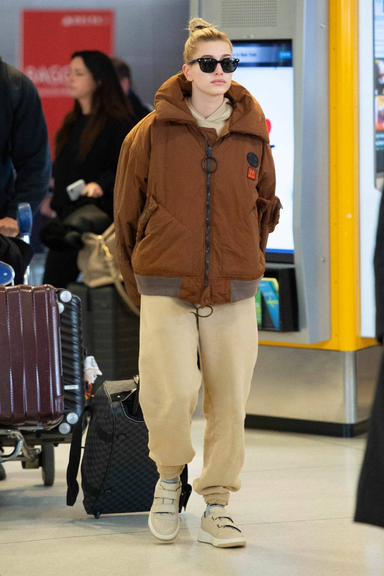 Hailey Baldwin-Biber på JFK i sportig jacka i brunt samt beige seatpants och hoodie under. Hon har solglasögon beiga sneakers och drar på en rullväska.
