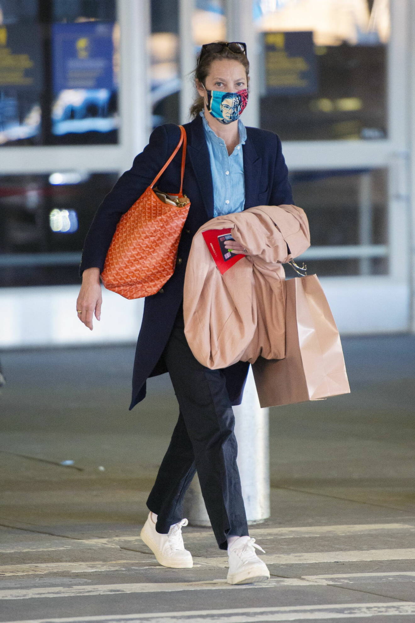 Christy Turlington i tunn rock, blå skjorta, svarta byxor och vita sneakers. På axeln har hon en orange shopper i skinn och åå armen bär hon en beige rock. Hon har även ett mönstrat munskydd.