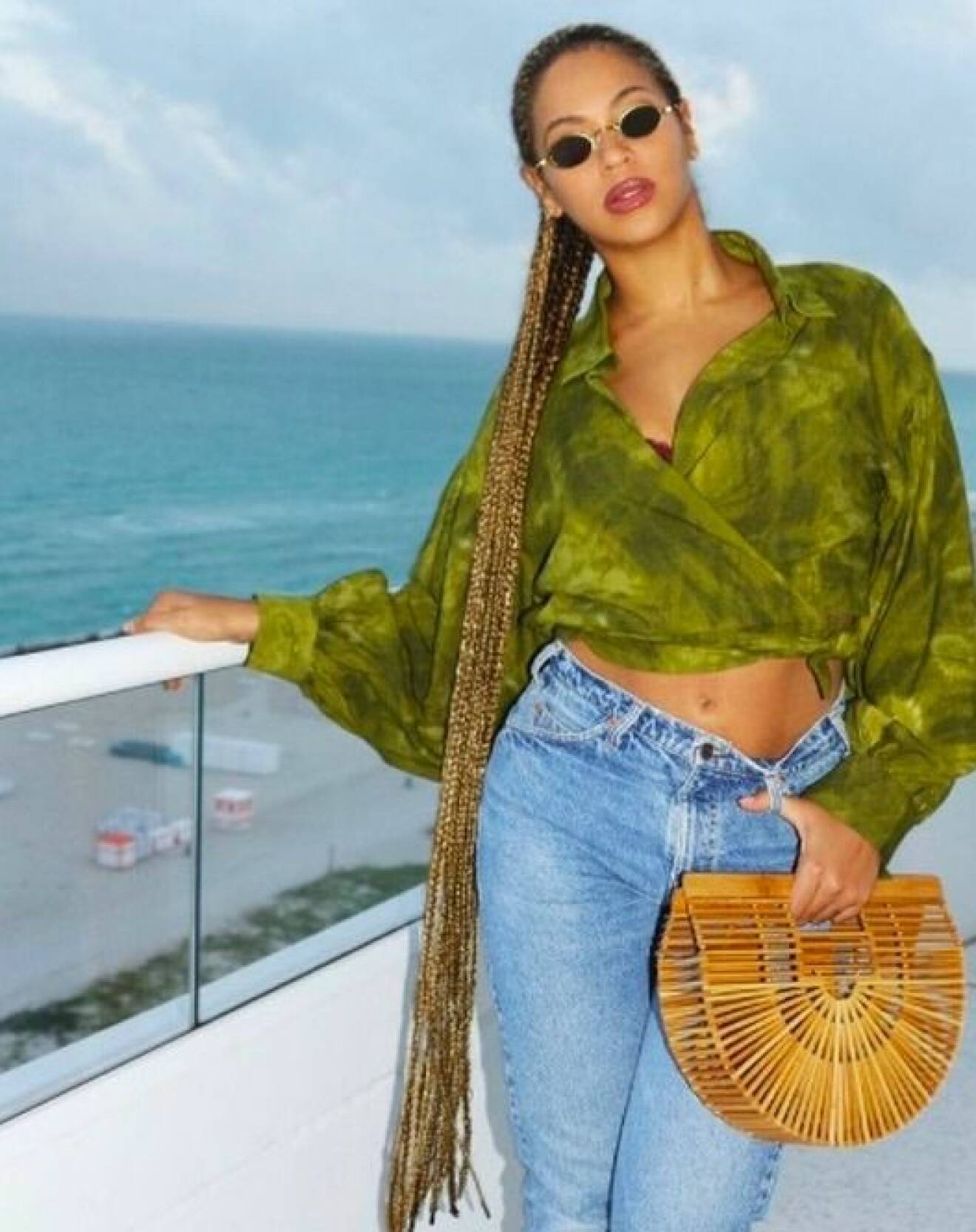 Beyoncé i grön batiksjorta och ljusblå jeans fotad på en balkong med en handväska i bambu. Väskan är formad som en halvcirkel och stel i formen. Handväska från Cult Gaia.