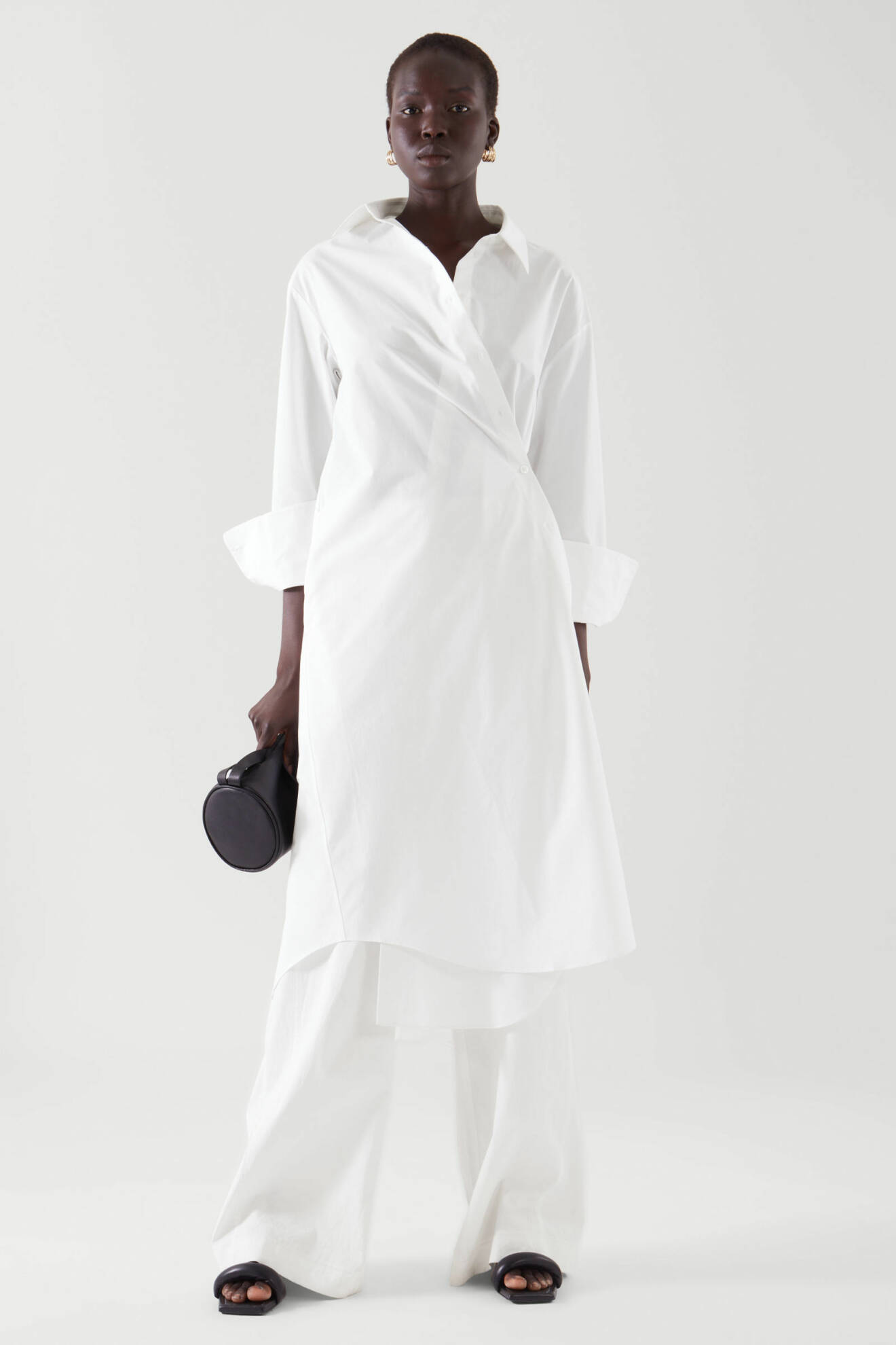 Modell med en lång vit skjortklänning som är omlott på kroppen. Går även att bära den som vanlig skjortklänning. Klänningen är stylad med vita raka byxor på bilden. Klänning från Cos.