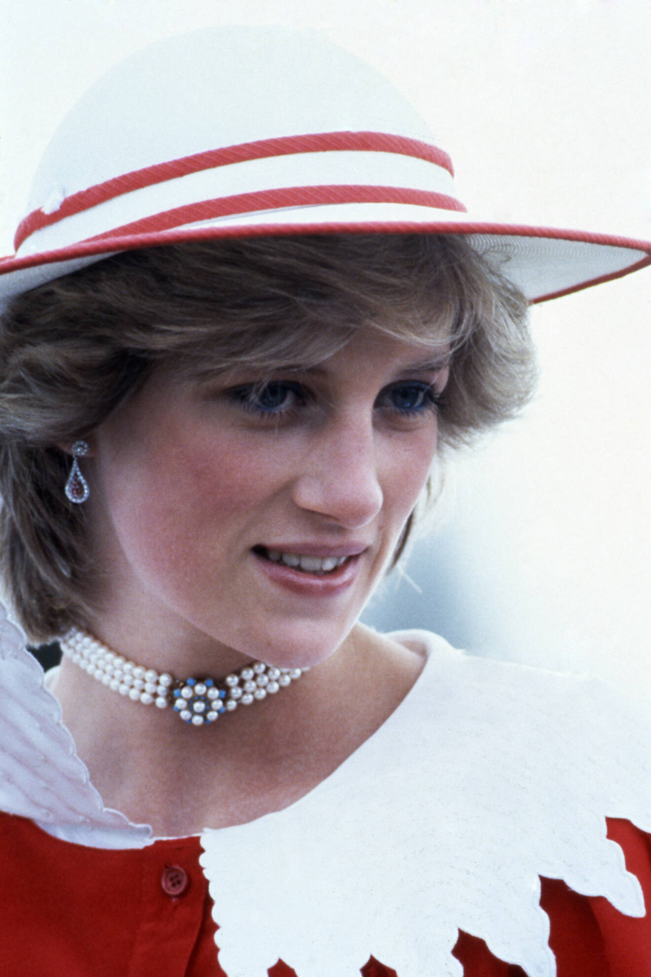 Prinsessan Diana i en röd blus med stor, vit krage på. Kragen är utskuren som en klassisk spetsduk och ger outfiten en romantisk touch. På huvudet har hon en röd och vitt hatt.