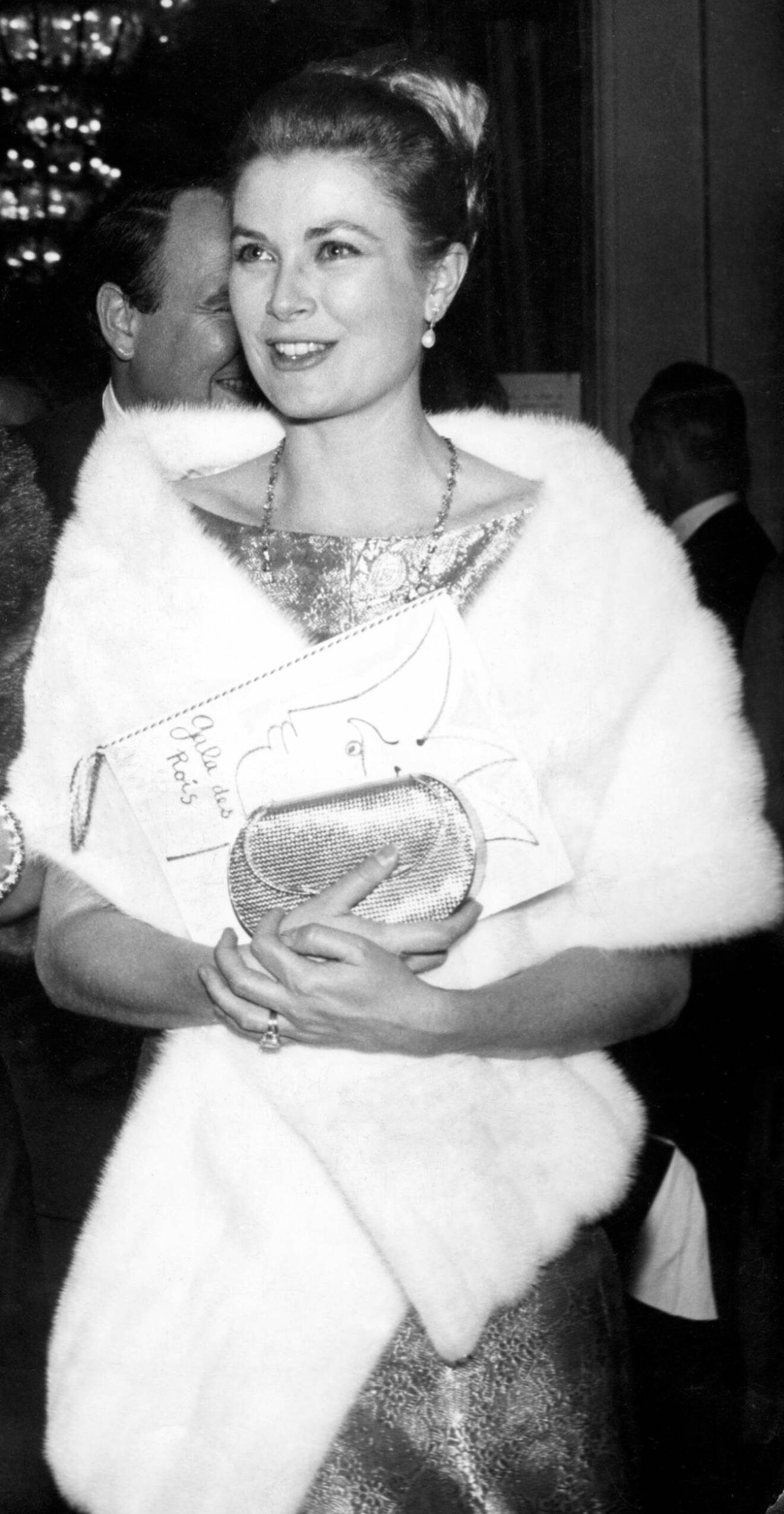 Grace Kelly i vit päls och uppsatt hår. Hon håller en bok och en kuvertväska i handen och man skymtar hennes stora förlovningsring på vänstra fingret.