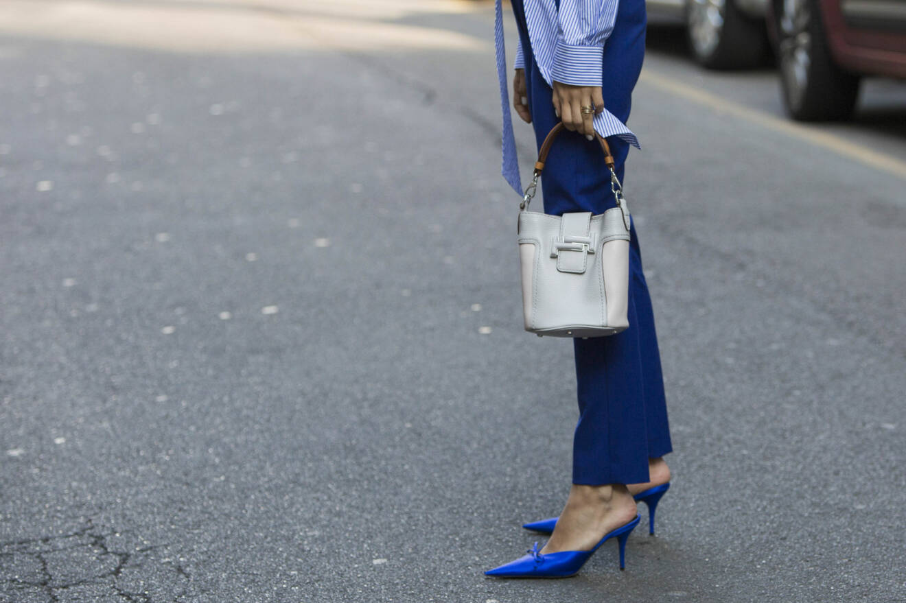 Närbild på en kvinna med blå byxor och blå pumps och en off-white skinnväska.