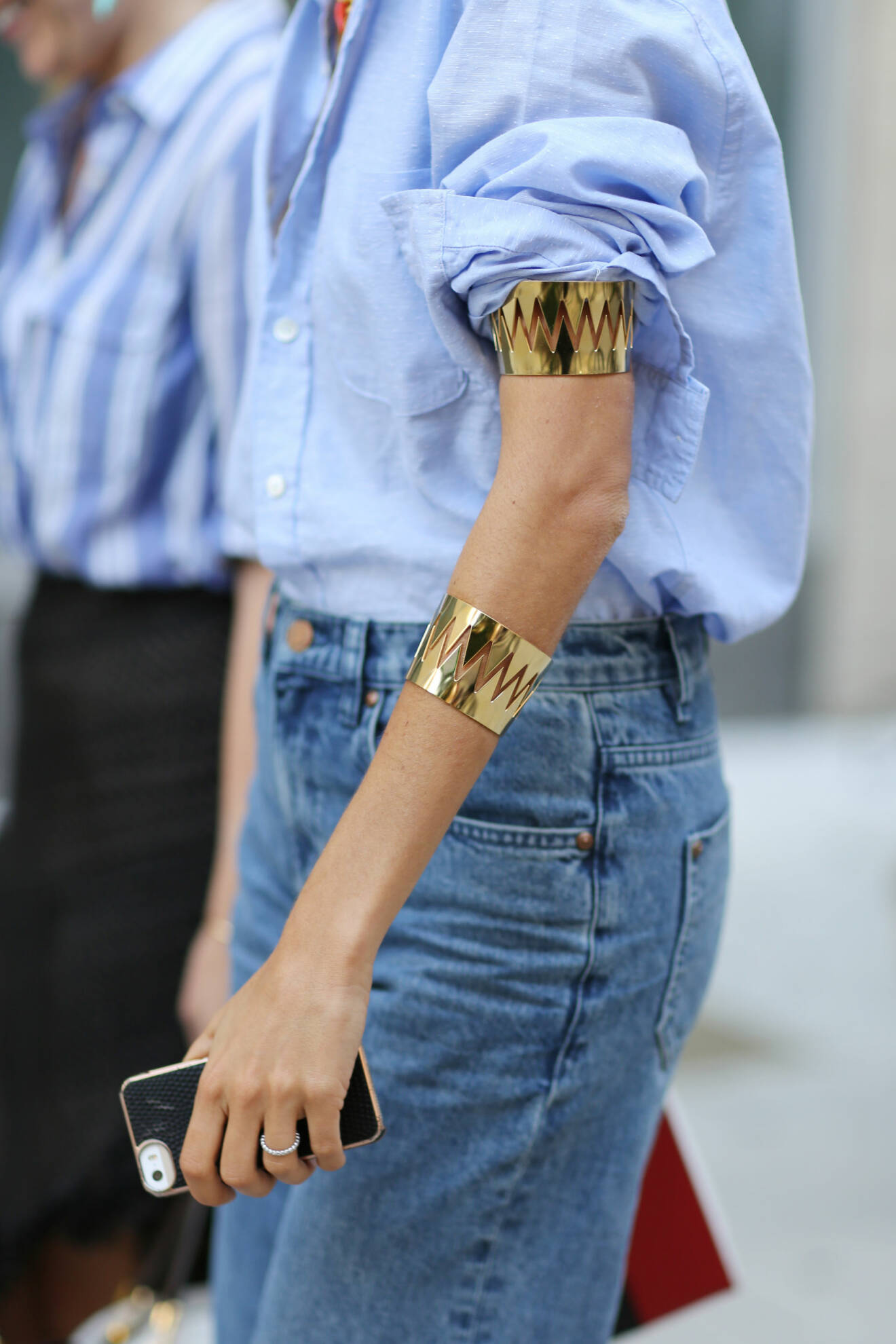 Närbild på kvinna i ljusblå skjorta och blå jeans och breda men enkla, gulfärgade armband och armring.