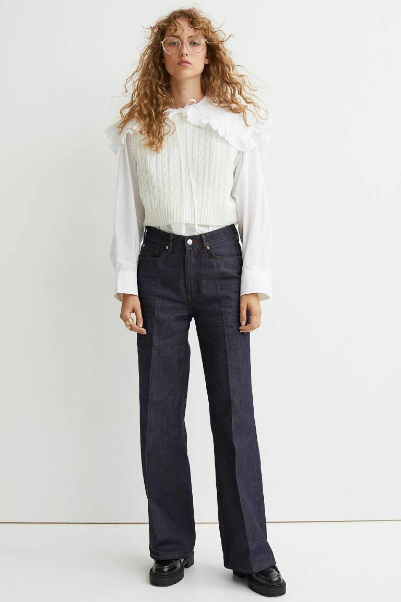 Modellen med mörka, blå, utställda jeans med pressveck. Till jeansen har hon en vit blus med stor, krage med volang och kabelstickad vit väst över. Outfit från H&amp;M.