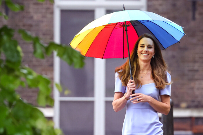 Kate Middleton i ljusblå klänning med ett färgglatt paraply i olika färger. Runt halsen bär hon tunna smycken med berlocker.