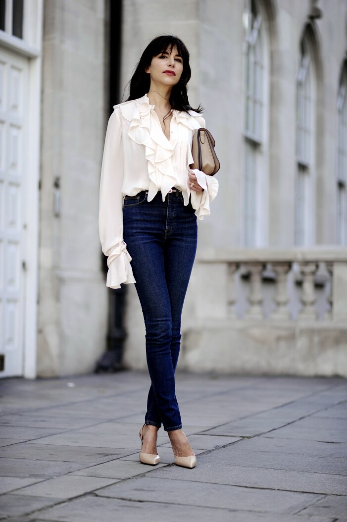 En skir blus och klassiska mörka jeans ger en lyxig look. Foto: TT. Kvinna i krämfärgad, transparent volangblus och mörkblå jeans utan slitningar. Hon har en beige väska och beiga, spetsiga pumps.