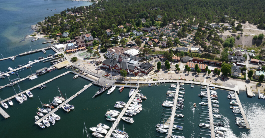 Drönarbild över Sandhamn med hamnen och KSSS seglarhotell i bakgrunden.Sedan slutet av 1800-talet har rika stockholmare haft semesterön Sandhamn som sitt sommarparadis. Sandhamn