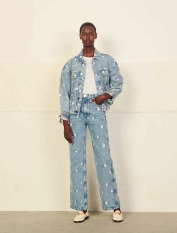 Outfit från Sandro. Modell med jeans och matchande jeansjacka i ljusblått med vita, broderade blommor på. Under jeansjackan har modellen en vit t-shirt och på fötterna vita loafers med guldfärgat spänne på.