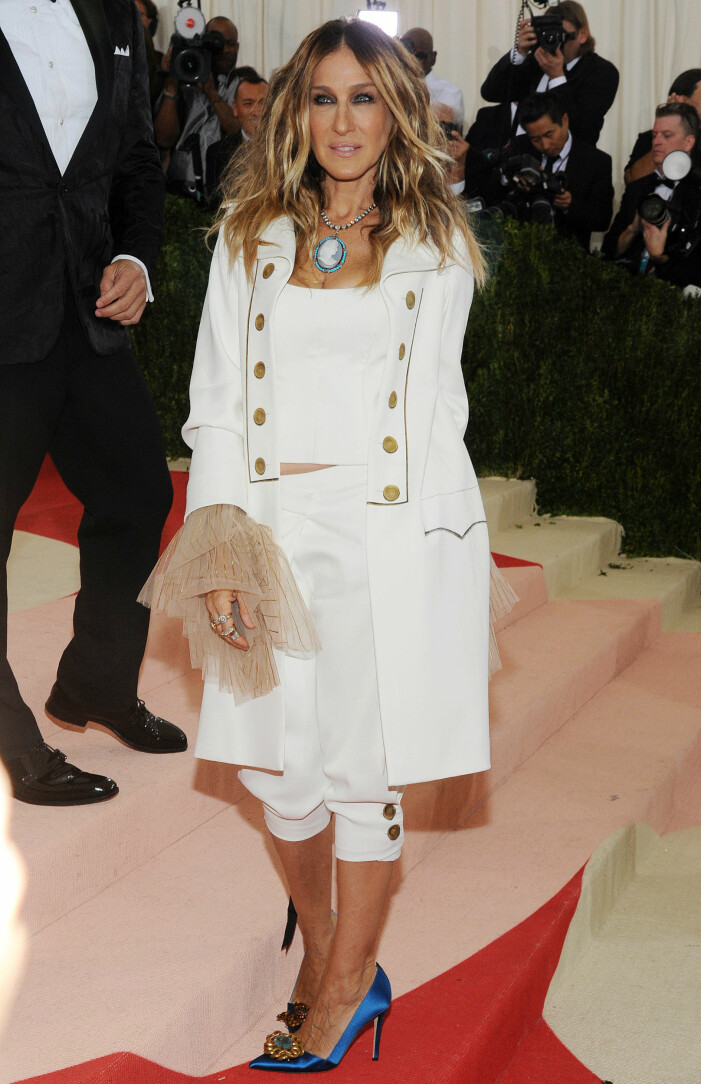 Sarah Jessica Parker tolkar temat "Teknologi" på Met-galan år 1996. Sarah Jessica Parker på Met-galan 1996 i vit outfit med kavajrock, linne och knälånga shorts. Hon har ett stort halsband och knallblå pumps med hög klack och spetsiga tår.