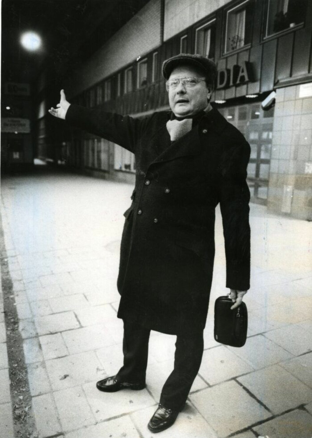 Den så kallade Skandiamannen, Stig Engström, i keps och med handledsväska fotograferad vid hans arbetsplats på Skandia på Tunnelgatan/Sveavägen. En man i svart rock och keps pekar med hela handen