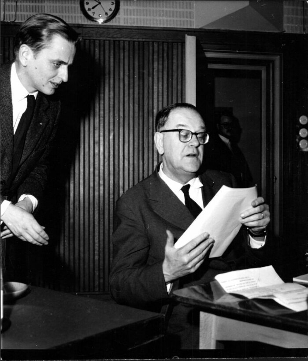 En bild på Tage Erlander, med en nyfiken Olof Palme bakom sig 1958. En man sitter vid ett skrivbord och tittar i papper, en annan man kikar fram över hans axel. Svart vit bild