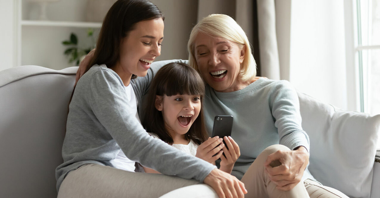 en äldre kvinna, en kvinna och ett barn sitter i en soffa och tittar i en mobil och skrattar
