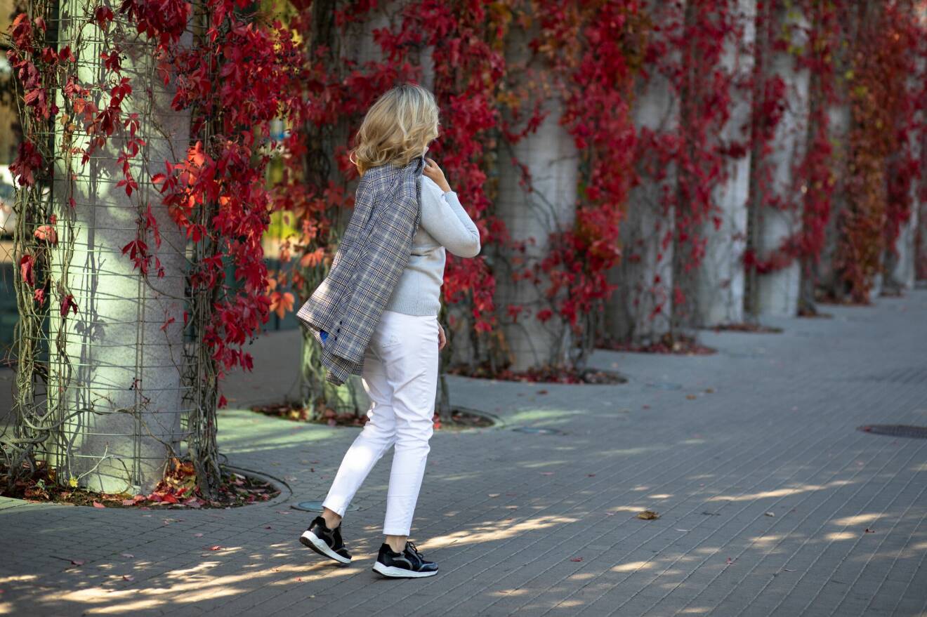 Kvinna med vita byxor och polotröja med sneakers och en kavaj över axeln under hösten.